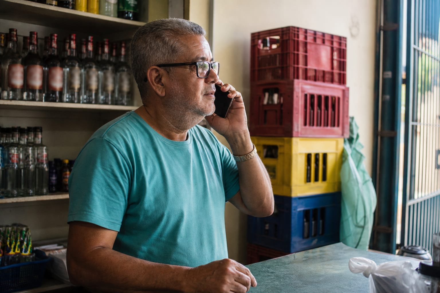 Dono de mercadinho de bairro usando o celular atrás do balcão com prateleiras de bebida ao fundo